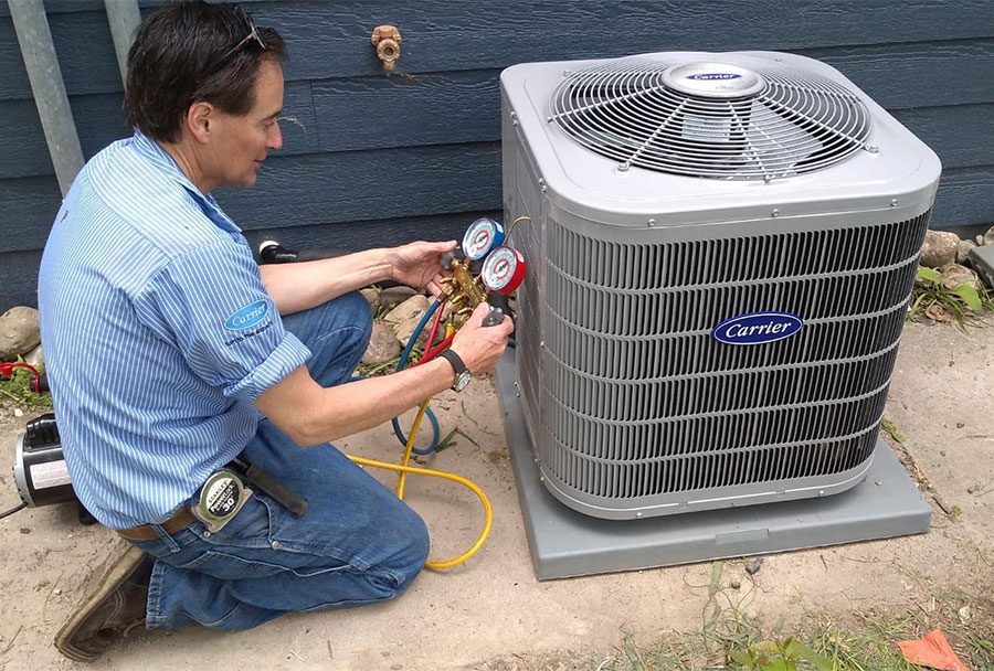 An AC repairman from Schneiss Heating & Air Conditioning in a blue shirt and jeans using  specialized tool on an AC unit in West Bend, WI